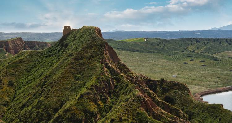 Grüner Hügel mit Graten und verfallener Steinturm mit Blick auf strukturiertes Ackerland und entfernte Berge.