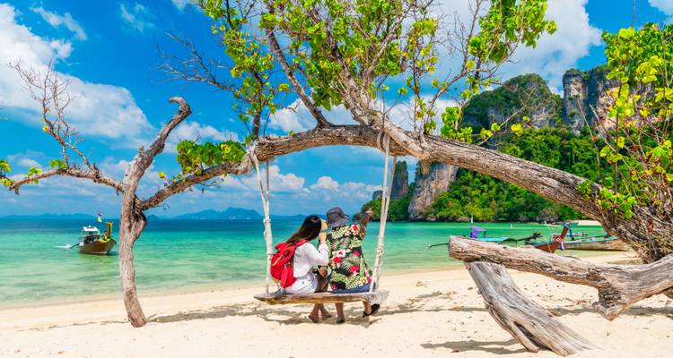 Couple relax on a wooden swing hung from a beach tree facing turquoise Andaman waters and long-tail boats.