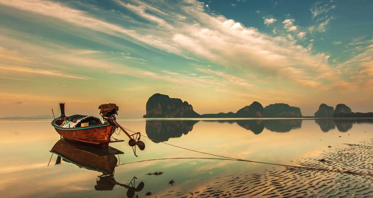 Long-tail boat moored on glassy water at sunrise with karst islands silhouetted on the horizon.