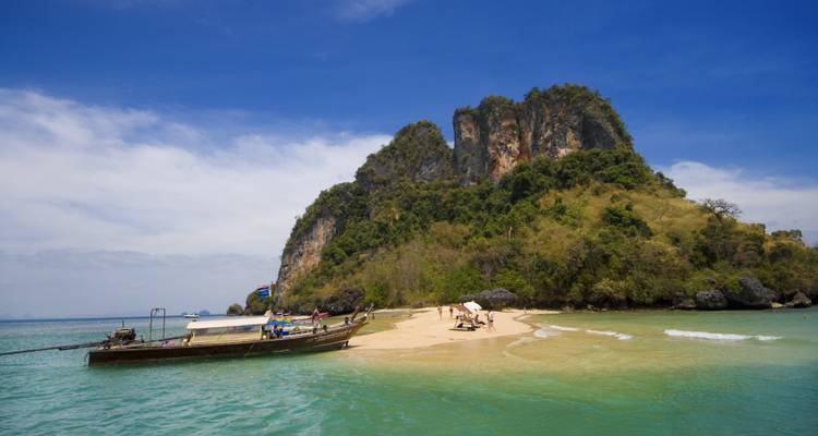 Traditional long-tail boat anchored at a tiny sandy islet beside a soaring limestone karst in turquoise waters.