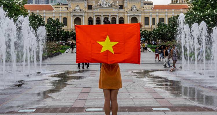 Persona sosteniendo una bandera vietnamita frente a fuentes con el ayuntamiento colonial desenfocado al fondo.