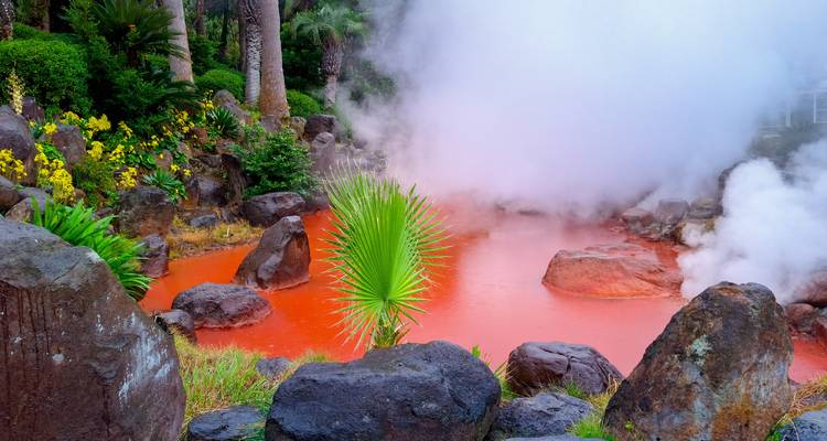 Vivid red geothermal hot spring surrounded by rocks, steam and tropical plants.