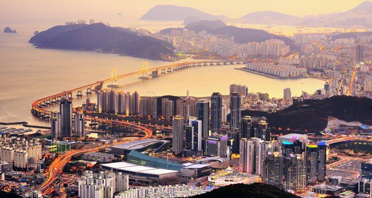 Sweeping dusk view over Busan with Gwangan Bridge lit up and mountains fading in the haze.