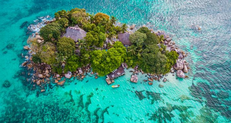 Vue aérienne d'une minuscule île privée entourée de récifs turquoise avec une végétation luxuriante et une petite jetée.
