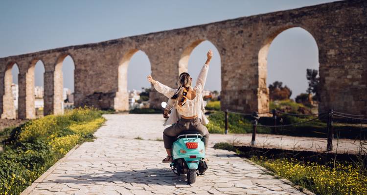 Two friends on a scooter raising their arms while passing ancient stone aqueduct arches.