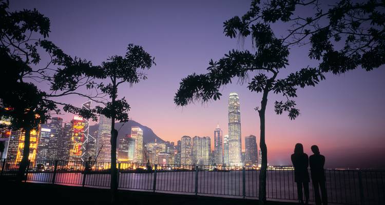 Silhouetted couple admire Hong Kong’s illuminated skyline at dusk framed by foliage.
