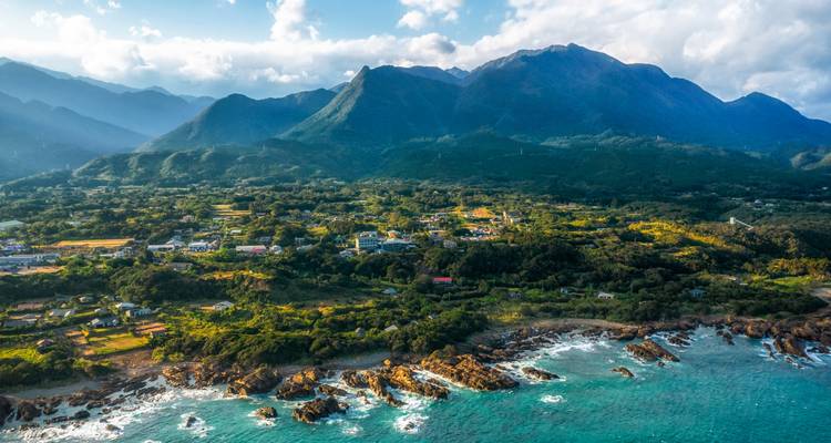 Aerial panorama of lush green island with rugged coastline and towering mountains rising behind a village.