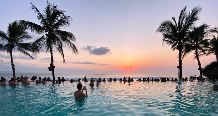 Crowd enjoys an infinity pool at sunset framed by palm trees overlooking the sea.