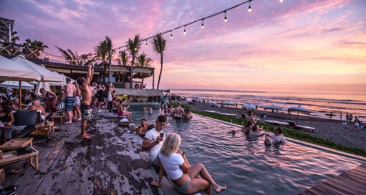 Lively beach club scene at dusk with people lounging in a pool and string lights overhead.