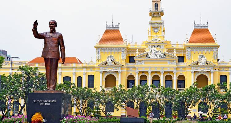 Statue of Ho Chi Minh waving in front of the ornate yellow colonial-era city hall in Ho Chi Minh City.