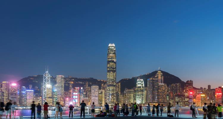 Nighttime city skyline with bright skyscraper lights and crowds gathered on a waterfront promenade.