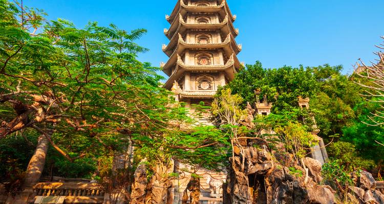 Tall pagoda framed by green foliage and rocky formations under a bright blue sky.