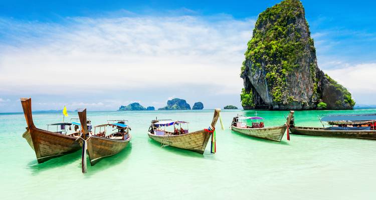 Row of colorful long-tail boats resting in shallow turquoise water with a dramatic limestone pinnacle beyond.