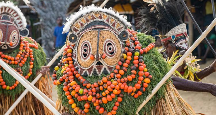Vibrantly dressed tribal performers in elaborate grass and bead costumes holding ceremonial poles.