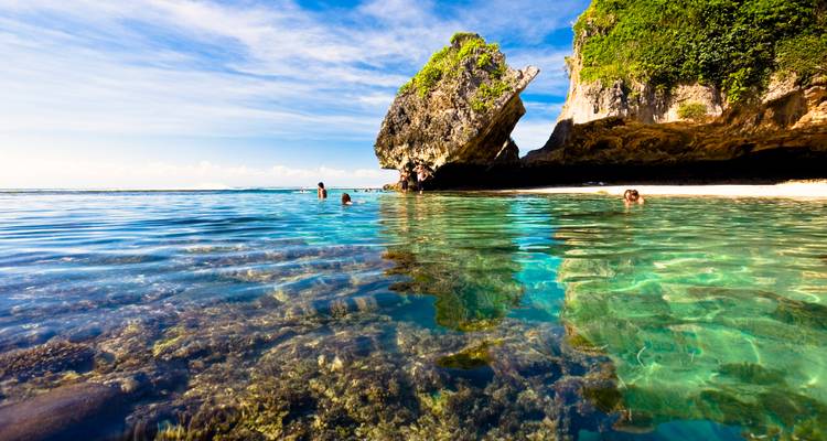 Swimmers enjoying crystal-clear turquoise water near a dramatic limestone outcrop on a Bali beach.