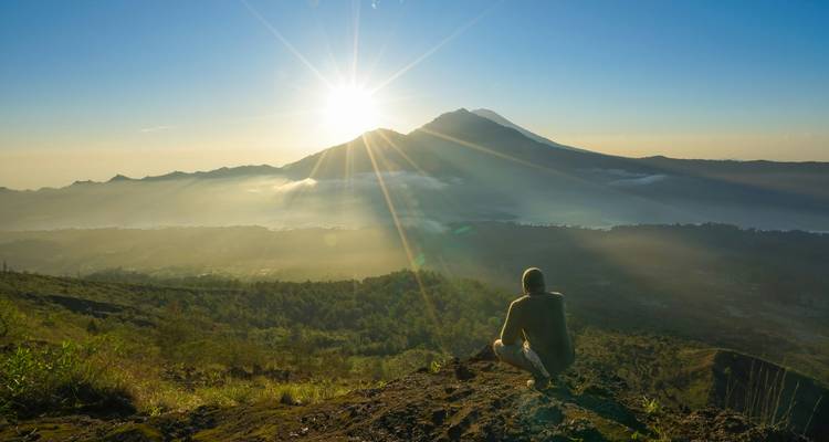 Hiker kneeling on a volcanic ridge watching sunrise over Mount Batur with sun rays spreading across the valley.