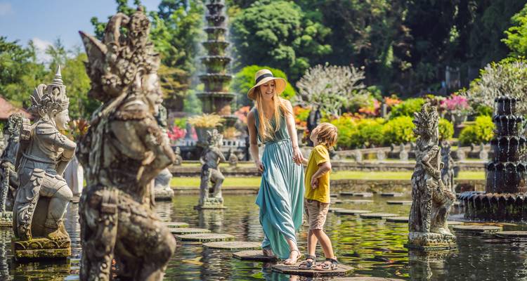 Mother and child crossing decorative stepping stones among ornate statues in a Balinese water palace garden.
