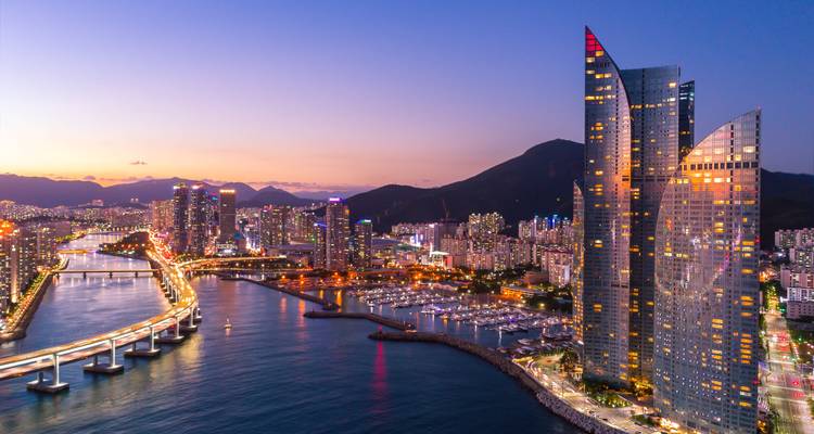 Twilight aerial of Busan’s skyscraper-lined marina and illuminated Gwangan Bridge curving over water.