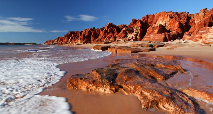 Wave-washed beach edged by striking red sandstone cliffs under a crisp blue Kimberley sky.