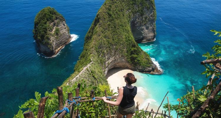 Young traveler stands at a cliffside railing looking down at a hidden white-sand beach and dramatic sea stacks.
