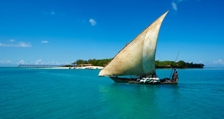 Velero tradicional dhow deslizándose sobre aguas azules tranquilas hacia una pequeña isla tropical.