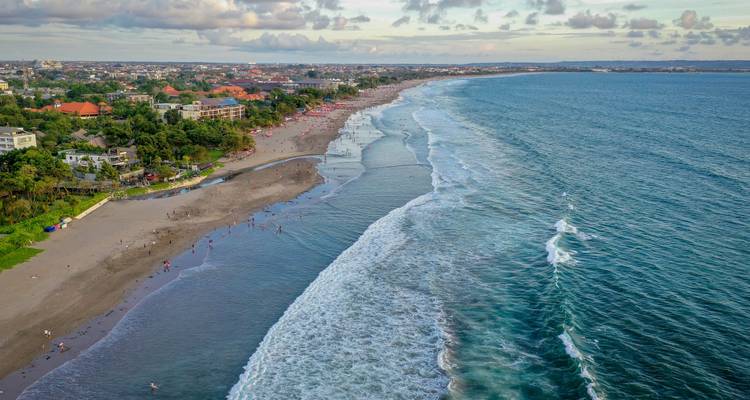 Aerial view of a long surf beach lined with resorts and breaking waves on a hazy afternoon.
