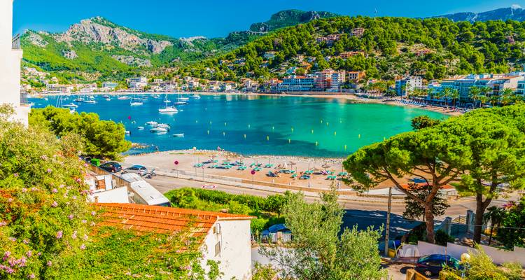 Bahía encantadora con agua color esmeralda, playa de arena y un pueblito colorido rodeado de montañas boscosas en Mallorca.