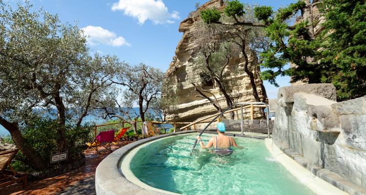 Woman enjoys a cliff-side thermal pool with sea views and olive trees surrounding.