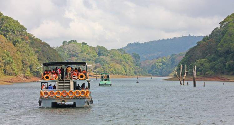 Tour boats cruise across Periyar Lake between forested hills under a cloudy sky.