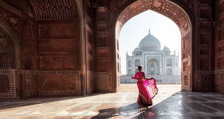 Woman in vibrant sari gazes at Taj Mahal through a grand Mughal arch at dawn.