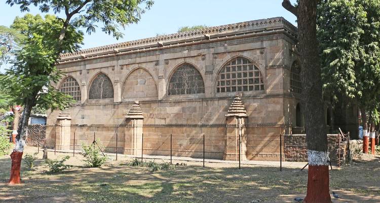 Simple stone tomb-like structure with arched windows set behind a fence amidst trees.