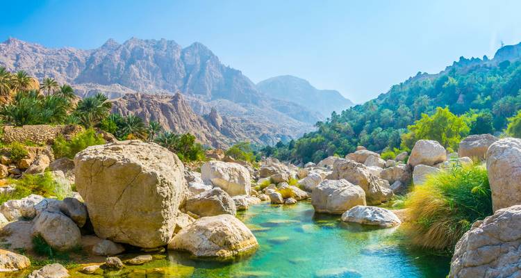 Lebendige türkisfarbene Becken und Felsbrocken in einem üppigen Canyon mit aufragenden Bergen des Wadi Shab.