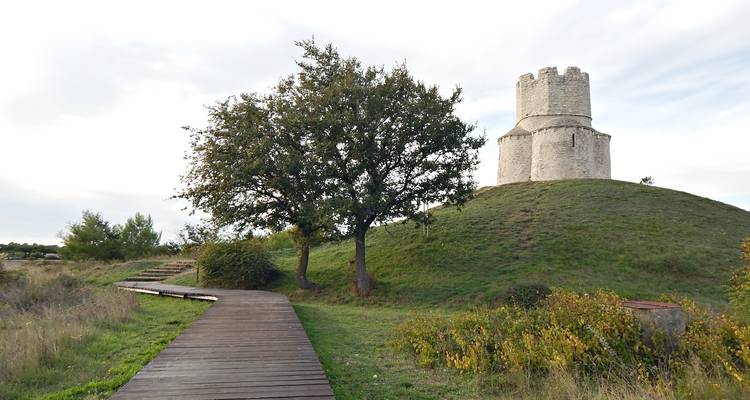 Una pequeña iglesia medieval corona una colina cubierta de pasto junto a un sendero de madera.