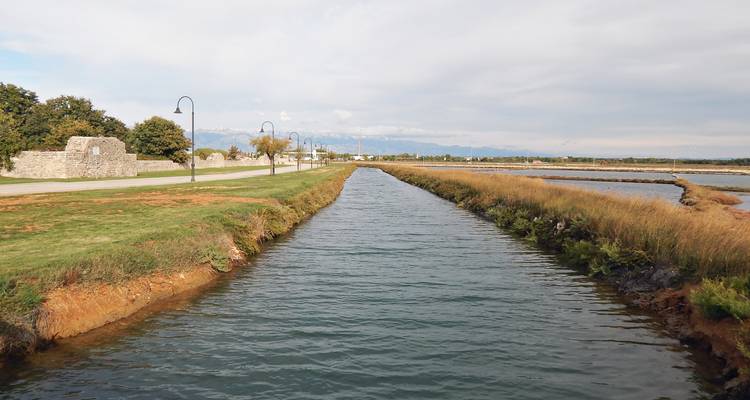 Un largo y estrecho canal flanqueado por orillas cubiertas de pasto que te lleva hacia montañas lejanas bajo un cielo brumoso.