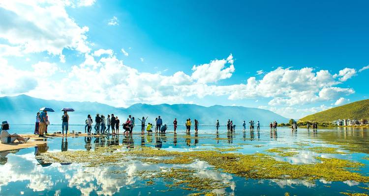Gente parada en un paseo marítimo de orilla de lago reflectante con montañas de fondo