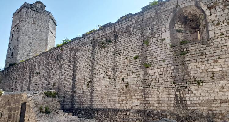 Tall medieval stone tower and lengthy defensive wall built from weathered blocks under a blue sky.