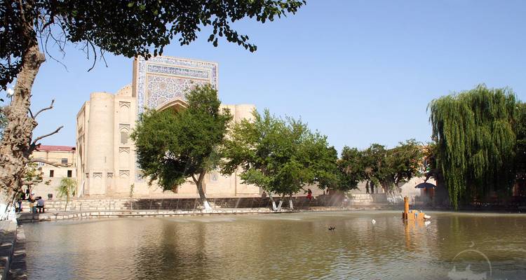 Serene pond framed by trees with a historic madrasa building reflected in the water.