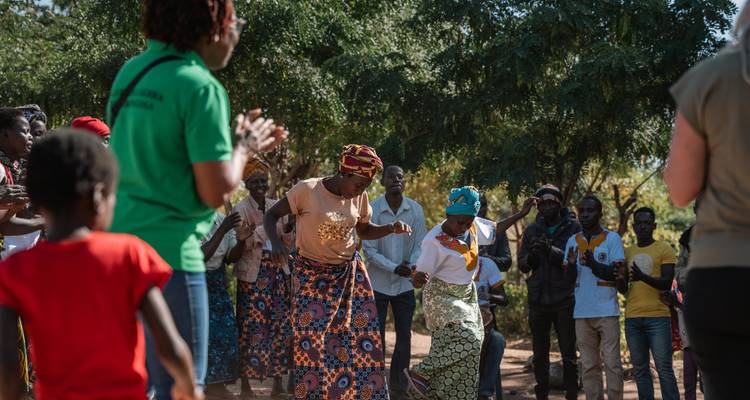 Local community members dancing outdoors while visitors look on and clap in a village setting.