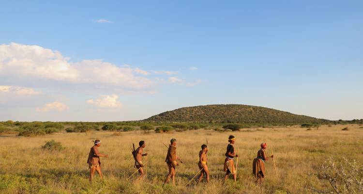 Line of San trackers walking through open grassland toward a distant hill under blue sky