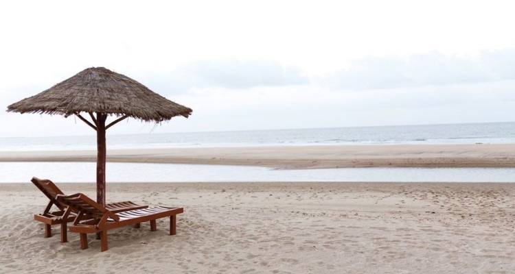 Two wooden loungers under a thatched umbrella on an empty beach by calm sea
