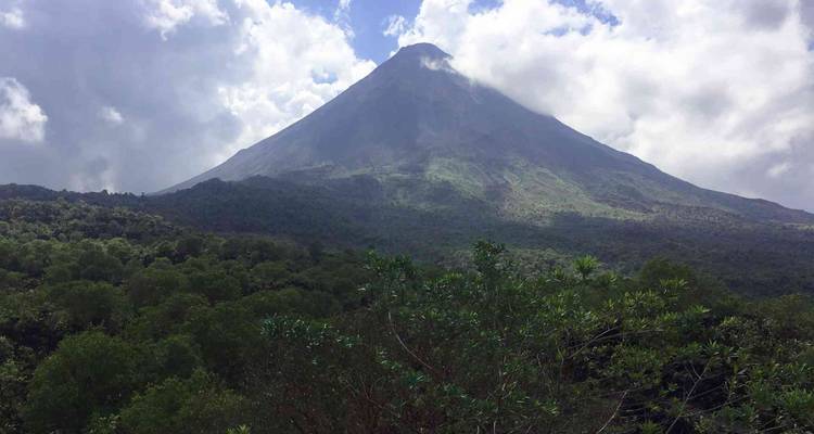 Volcan majestueux de forme conique s'élevant au-dessus d'une forêt tropicale dense sous un ciel partiellement nuageux.