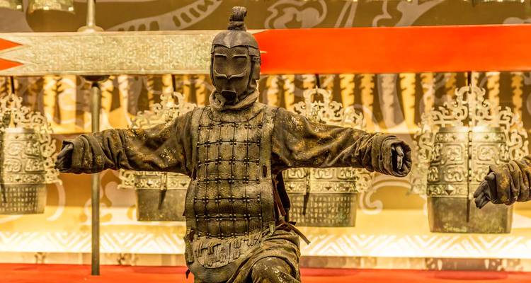 Armored terracotta warrior figure displayed amid ancient bronze bells