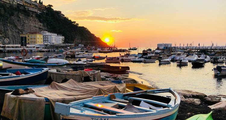 Small fishing boats rest in a harbor as the sun sets behind seaside cliffs.