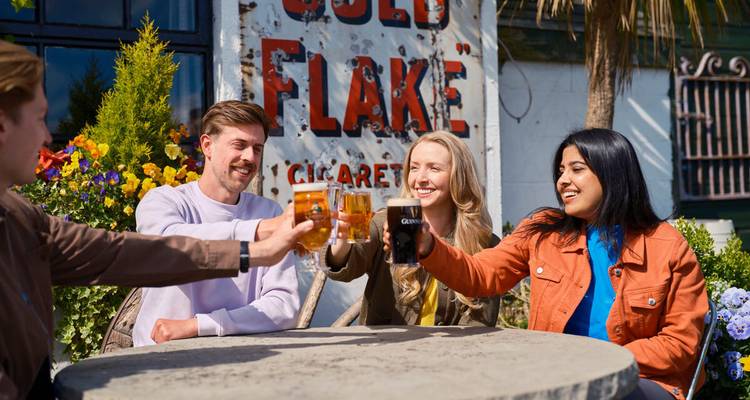 Grupo de amigos levantando jarras de cerveza en la terraza de un pub irlandés al aire libre rodeada de flores y señalética vintage