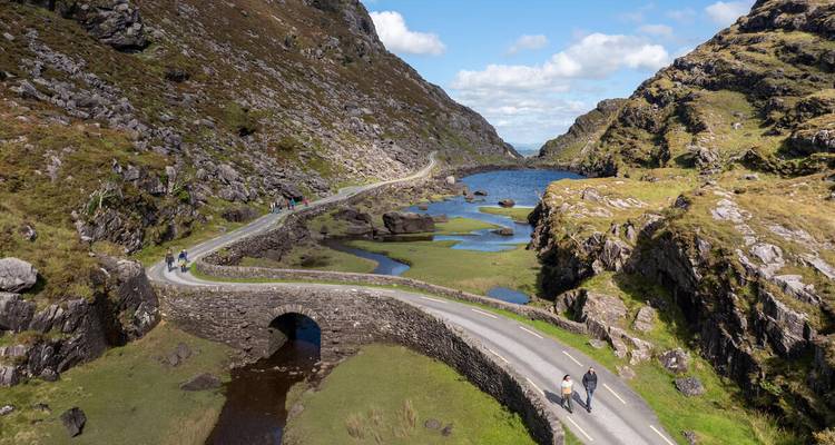 Vista aérea del sinuoso camino Gap of Dunloe que cruza un arroyo entre montañas verdes y escarpadas bajo cielos azules
