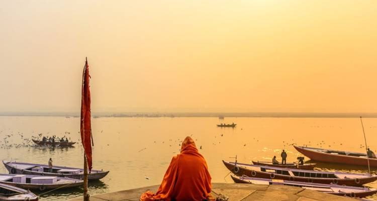 A lone monk draped in orange sits on the ghats facing the Ganges River at dawn, with wooden boats and a golden hazy sky.