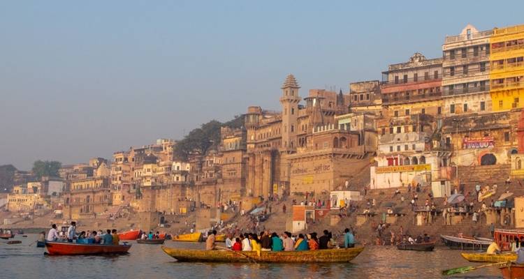 Colorful boats filled with pilgrims pass the riverside temples and sandstone buildings of Varanasi at dusk.