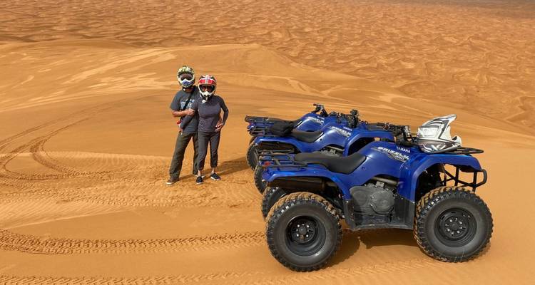 Two travellers in helmets stand beside blue quad bikes on vast orange sand dunes under clear skies, tyre tracks visible in the soft sand.