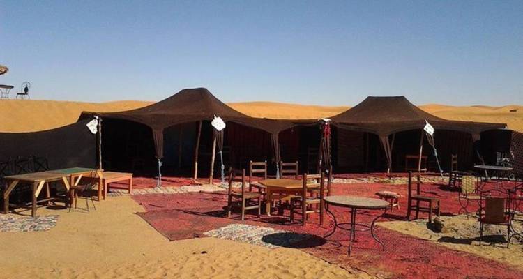 Simple desert camp with three dark canvas tents, scattered wooden tables and chairs on patterned rugs amid rolling golden dunes.