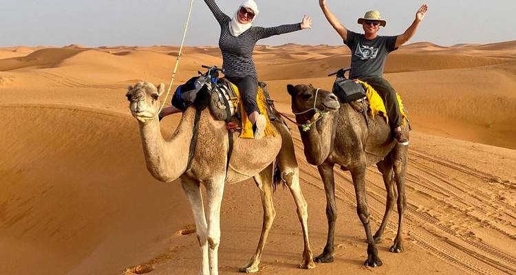 Two smiling travellers ride camels across sweeping orange Sahara dunes, arms raised in excitement, with clear blue sky overhead.